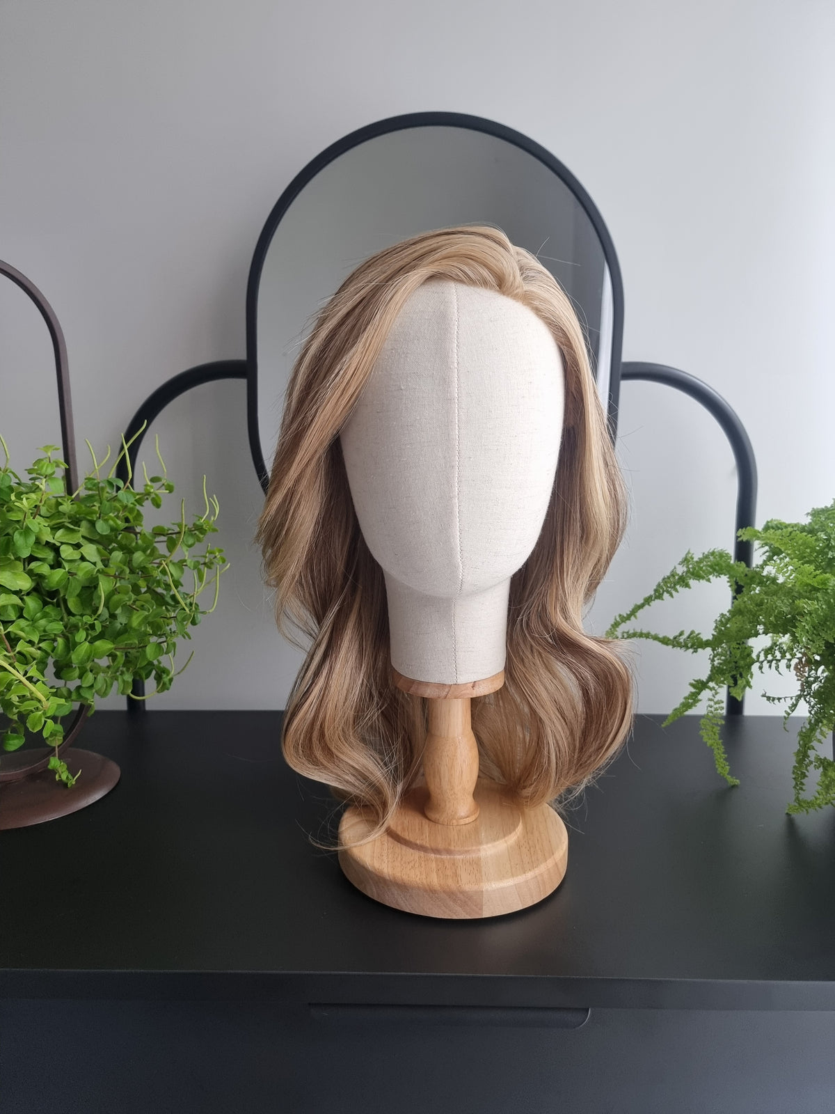 A shoulder-length wavy wig with blonde and light brown highlights, displayed on a cream fabric mannequin head mounted on a wooden stand, set on a black shelf with green plants and a decorative mirror in the background.