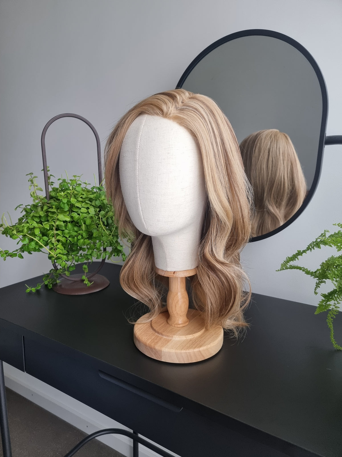 A shoulder-length wavy blonde wig displayed on a white mannequin head stand with a wooden base, set on a black table with a mirror, plants, and a gray wall in the background.