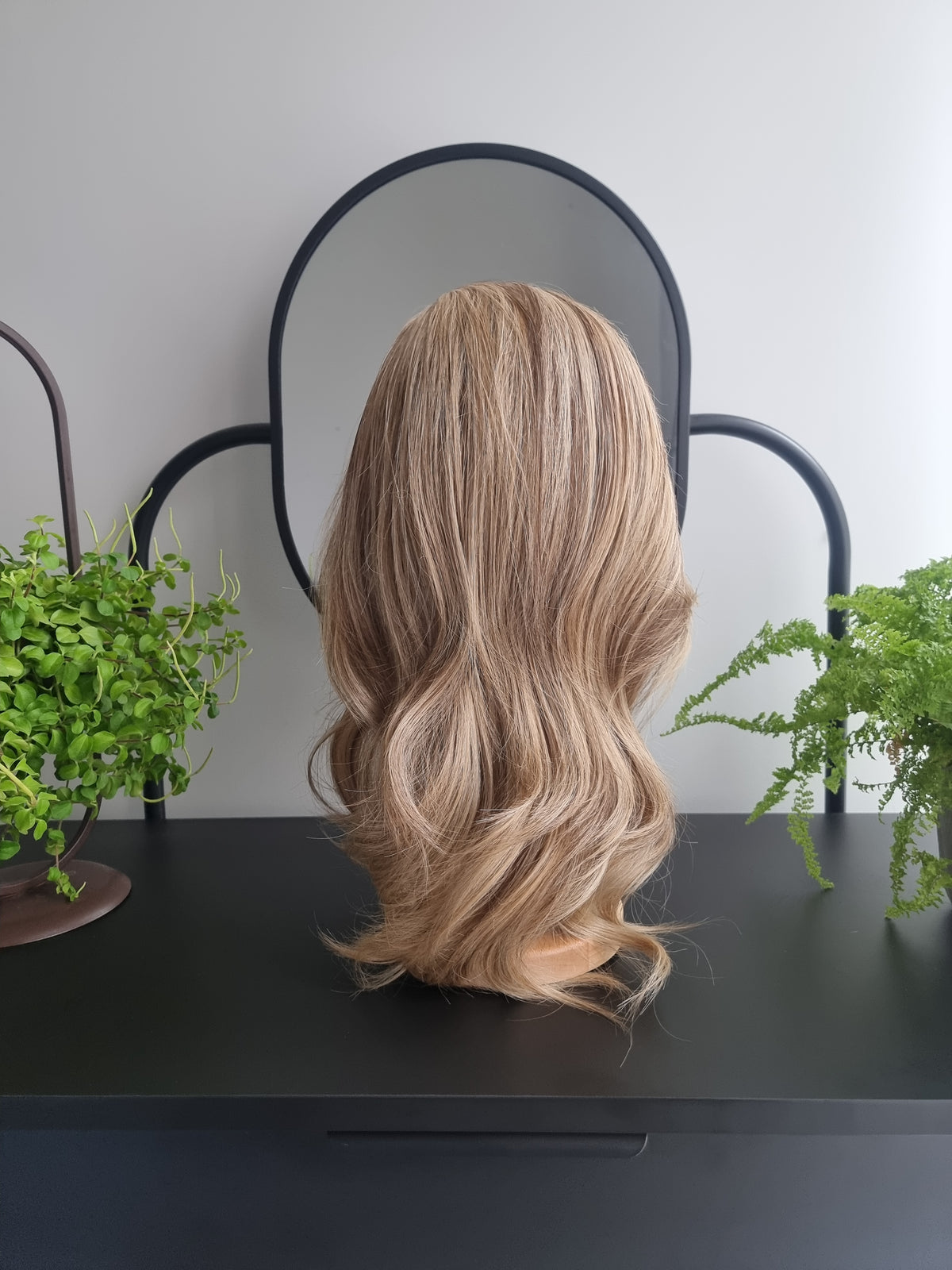 A shoulder-length wavy wig with blonde and light brown highlights, displayed on a mannequin head from the back, set against a minimalist background with a black table and mirror.