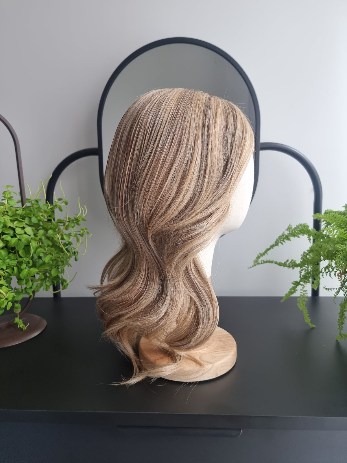 A shoulder-length wavy wig with blonde and brown highlights, displayed on a mannequin head stand on a black table with green plants in the background.