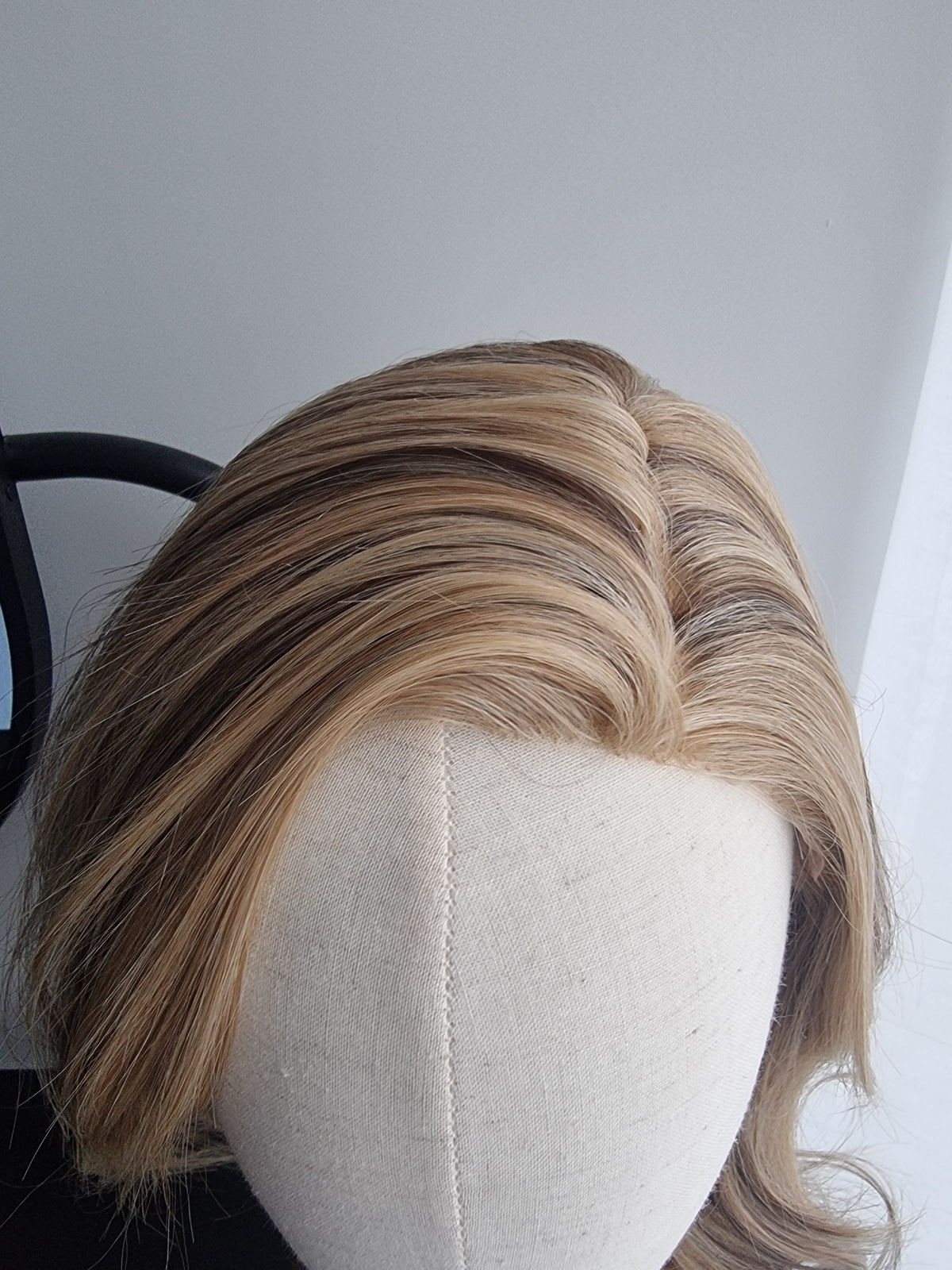 A close-up of a blonde wig with brown lowlights, styled on a cream-colored mannequin head against a plain light gray background.