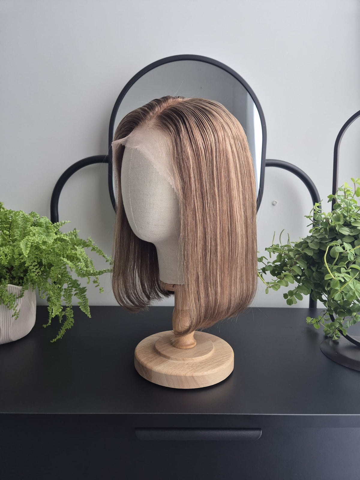 A shoulder-length straight wig with highlighted brown and blonde tones displayed on a beige mannequin head stand with a wooden base, set on a black table with potted green plants and a black-framed mirror in the background.