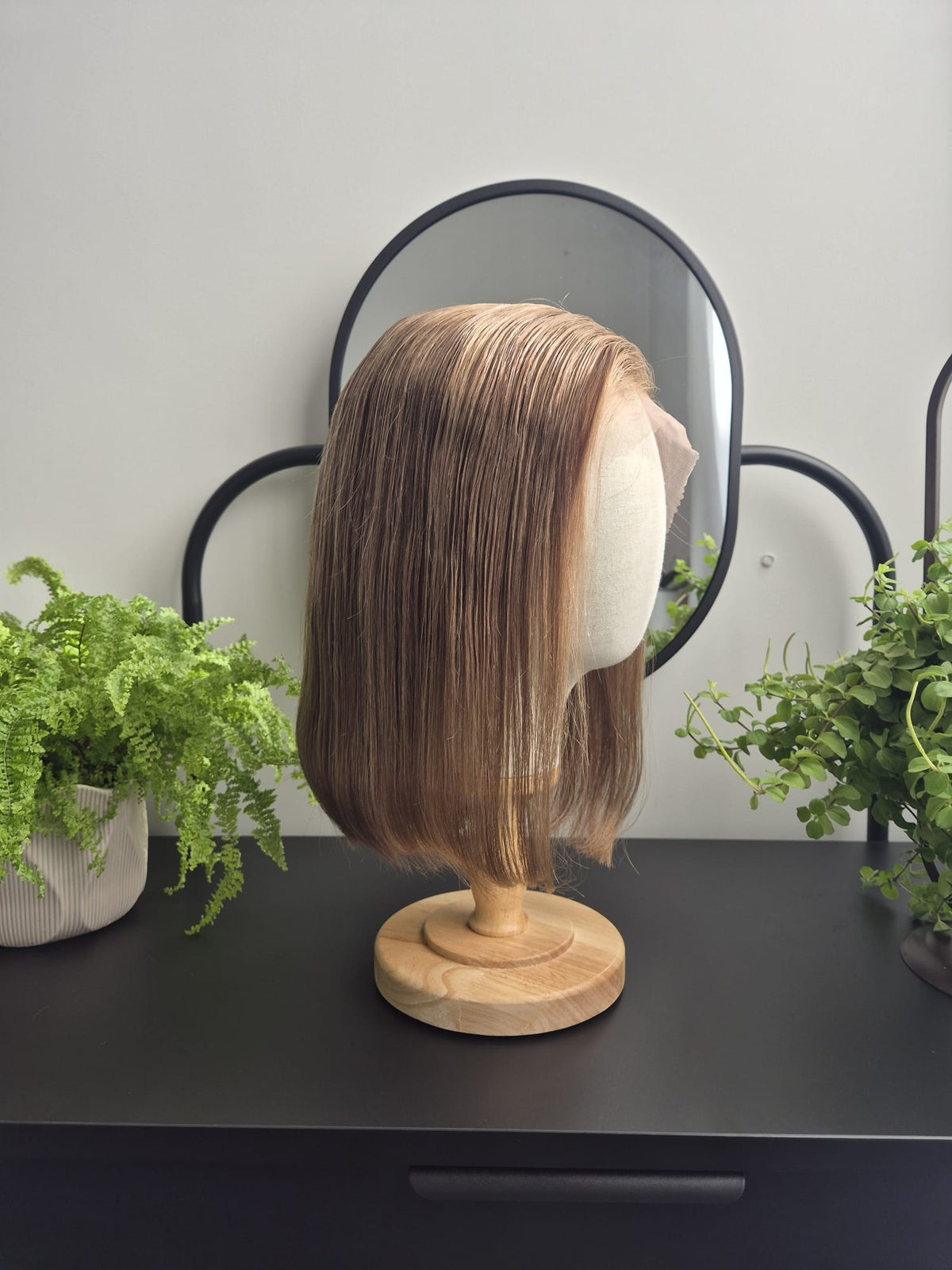 A shoulder-length straight wig with light brown to blonde highlights, displayed on a wooden stand with a mannequin head. The stand is placed on a black table with green potted plants and a black-framed mirror in the background.