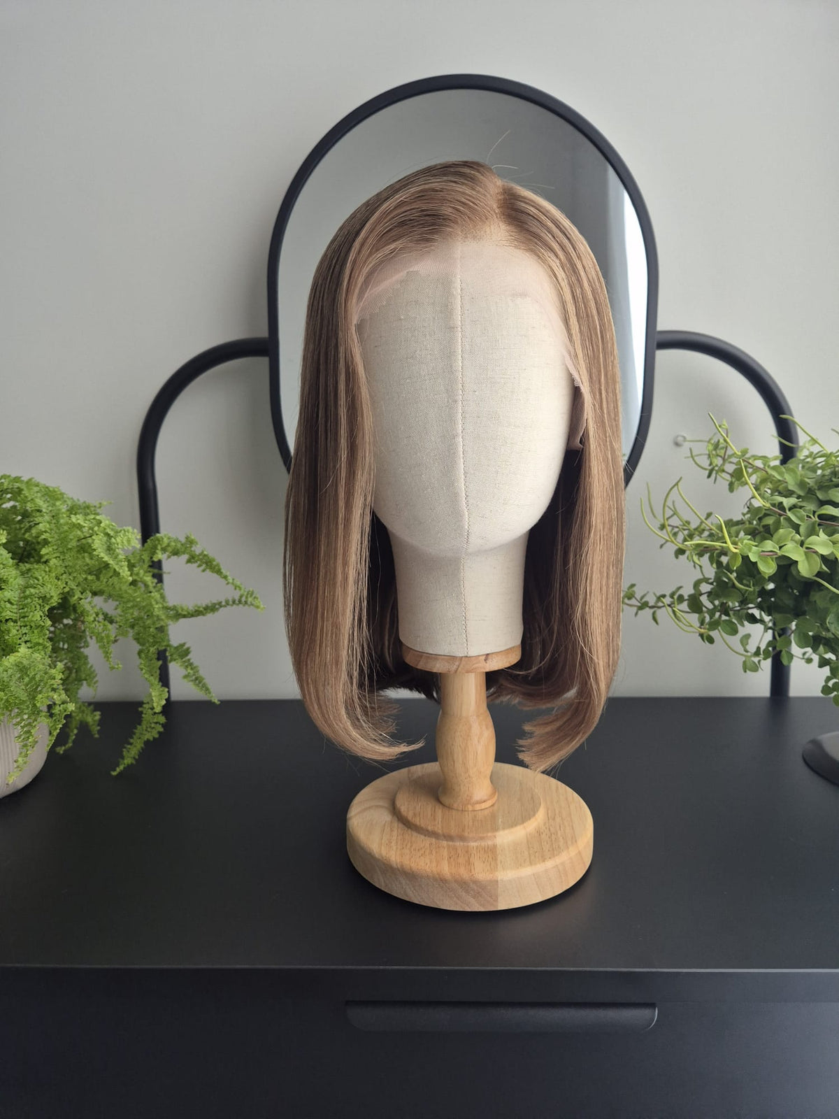 A shoulder-length straight wig with light brown to blonde highlights, displayed on a beige fabric mannequin head mounted on a wooden stand, set against a black-framed mirror on a dark table with green plants.